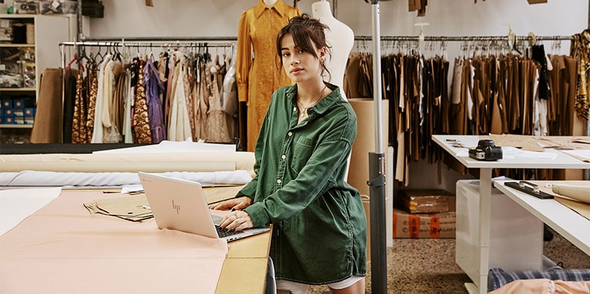 A woman working on some close designs on her HP laptop while standing in front of a wooden desk.