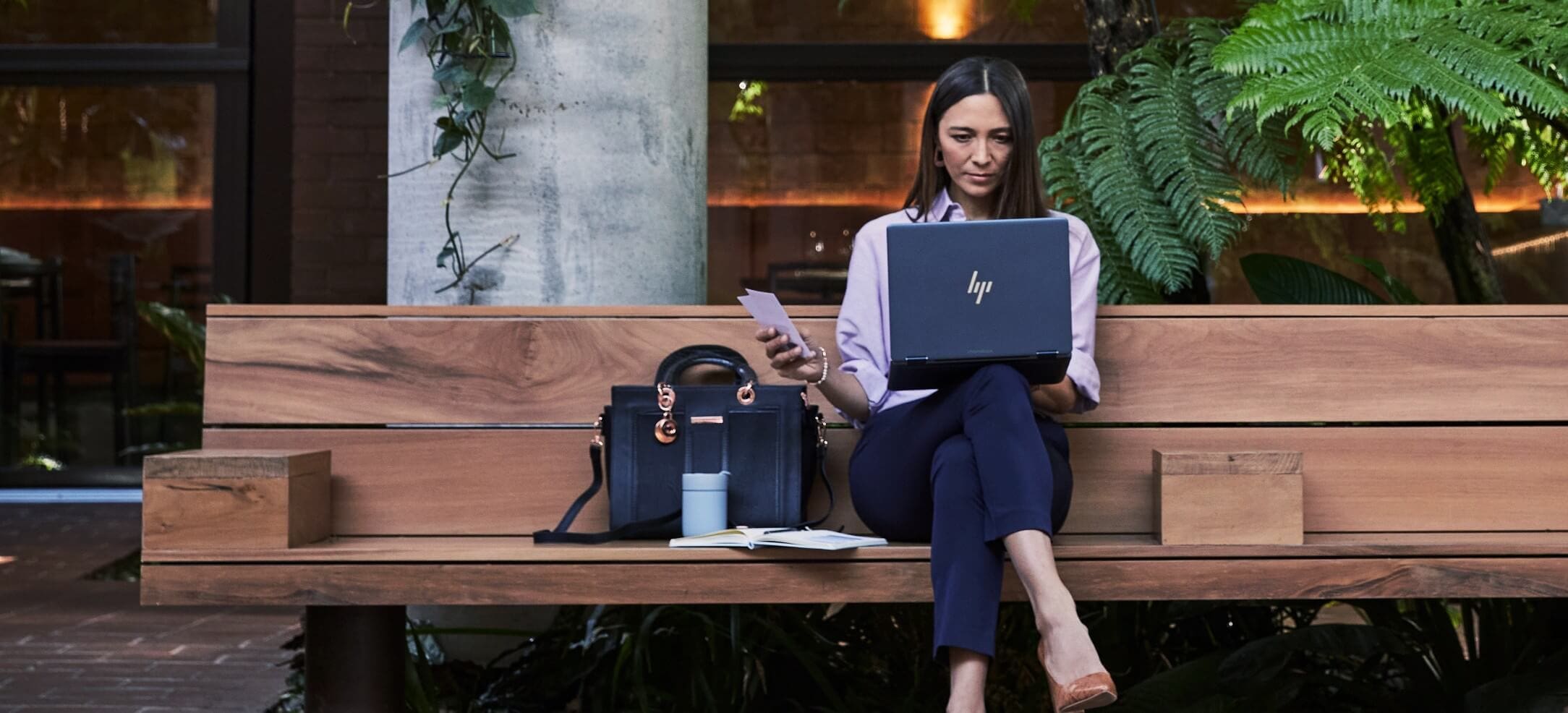 A woman sitting on a wooden bench while checking some data on her HP laptop on top of her lap.