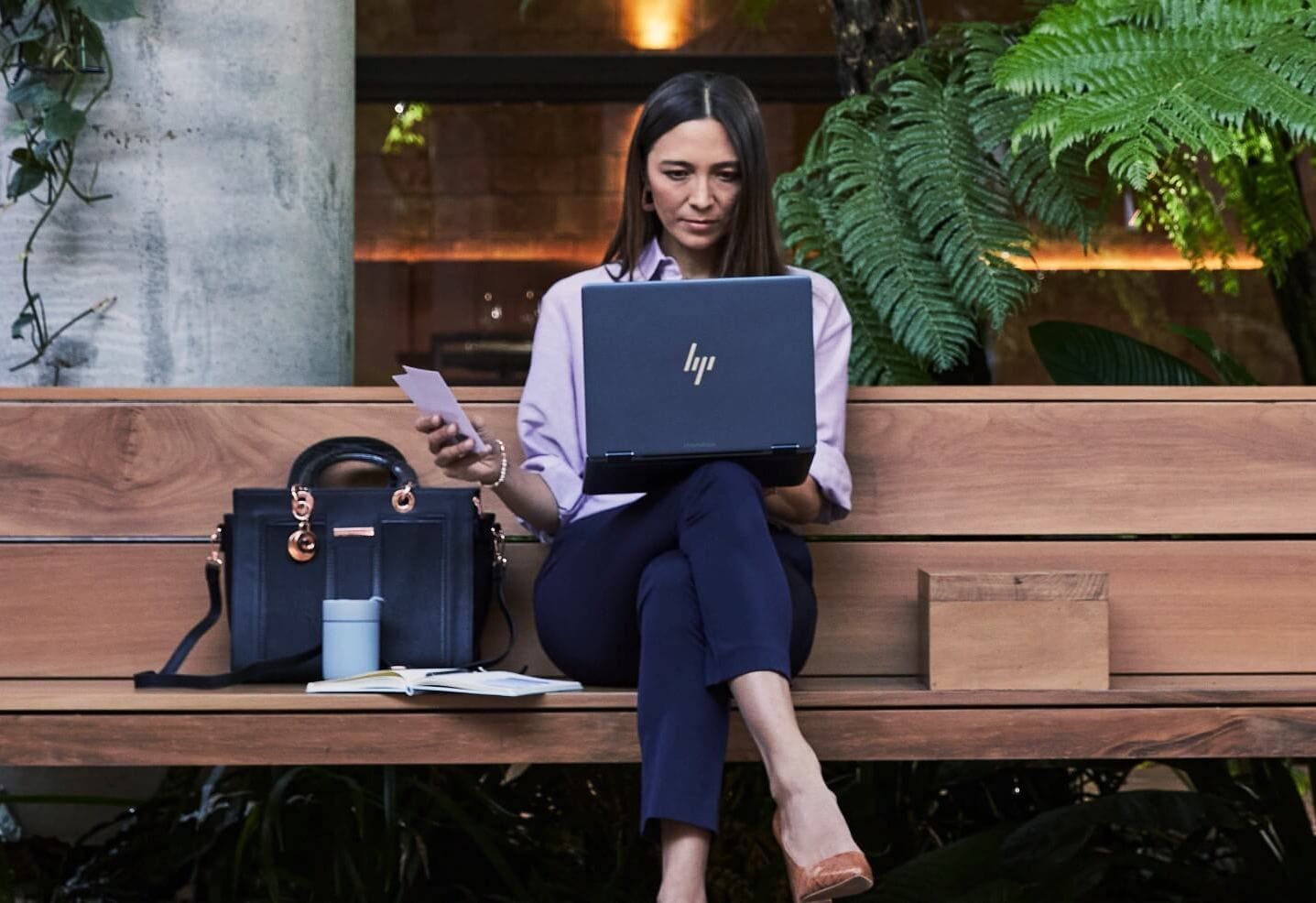 A woman sitting on a wooden bench while checking some data on her HP laptop on top of her lap.