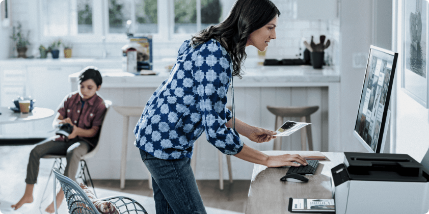 A woman holding a printout while looking at her HP monitor at home.