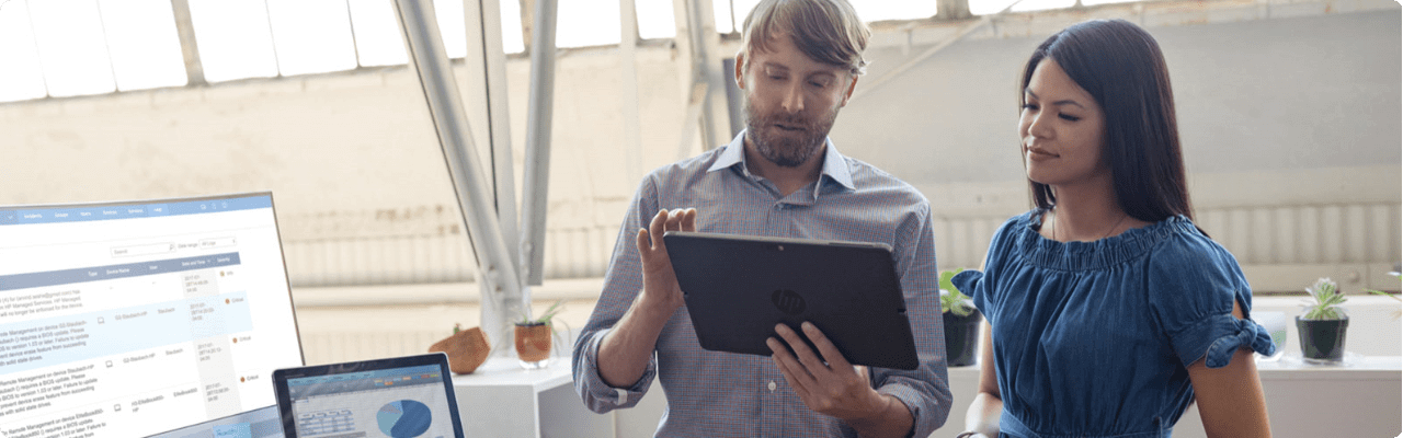 A man showing some data to his co-worker on his HP tablet while standing in front of his desk.