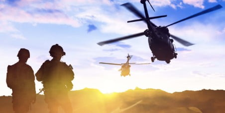 Two soldiers looking at two choppers flying in the desert