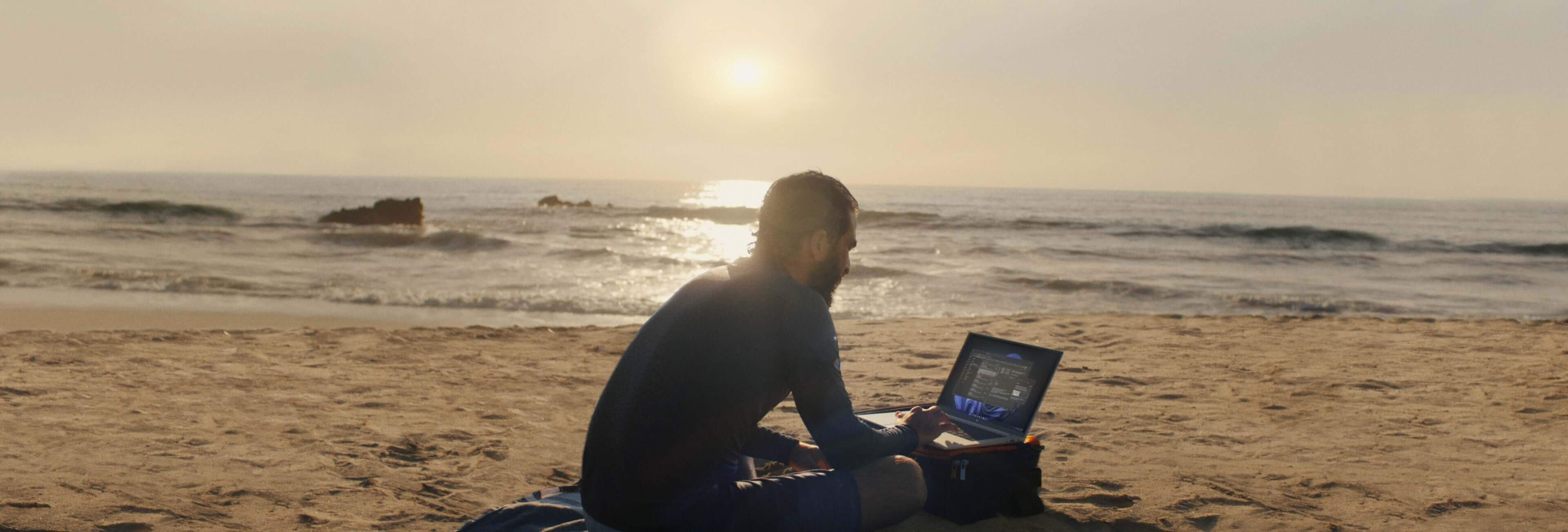 A man looking at some data on his HP laptop while sitting on a beach in the sunset.