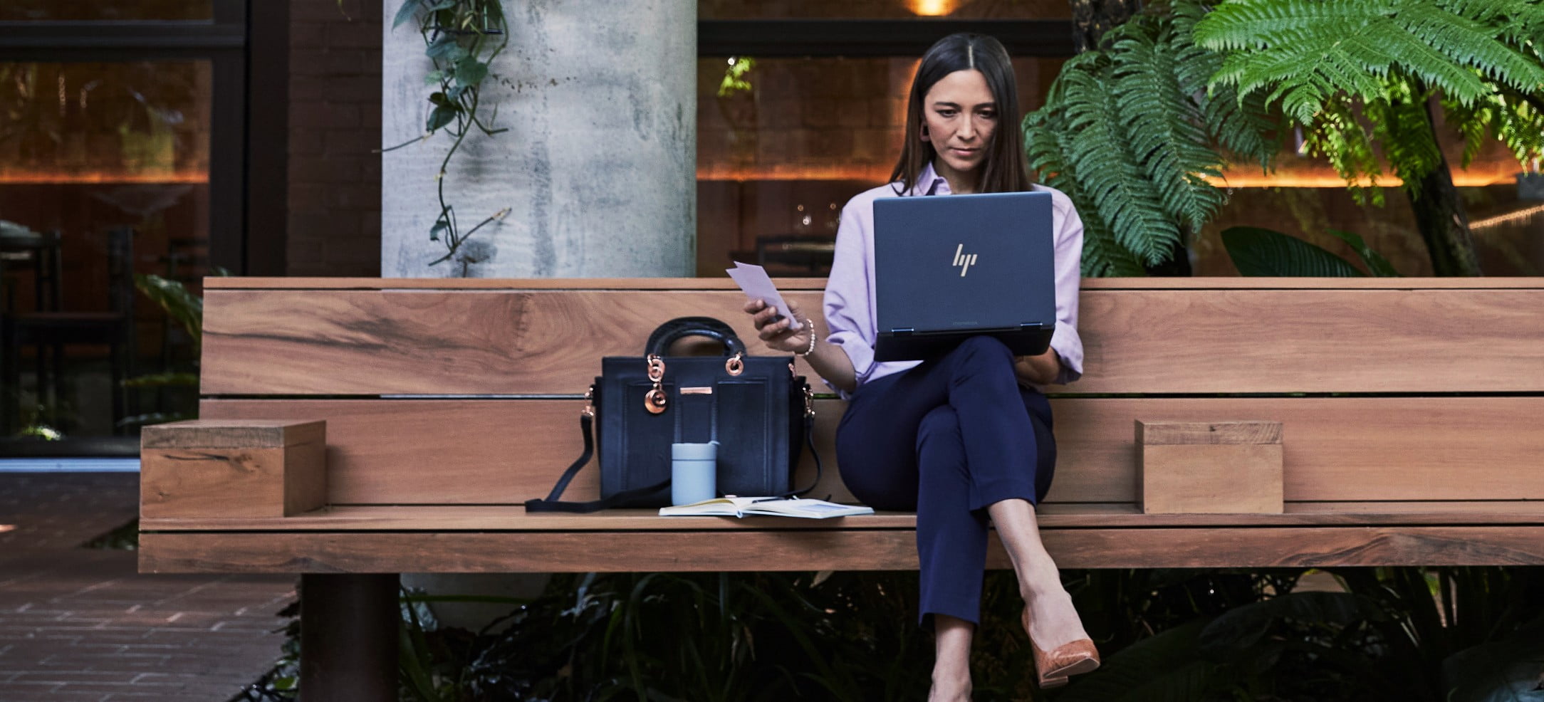 A woman sitting on a wooden bench while checking some data on her HP laptop on top of her lap.