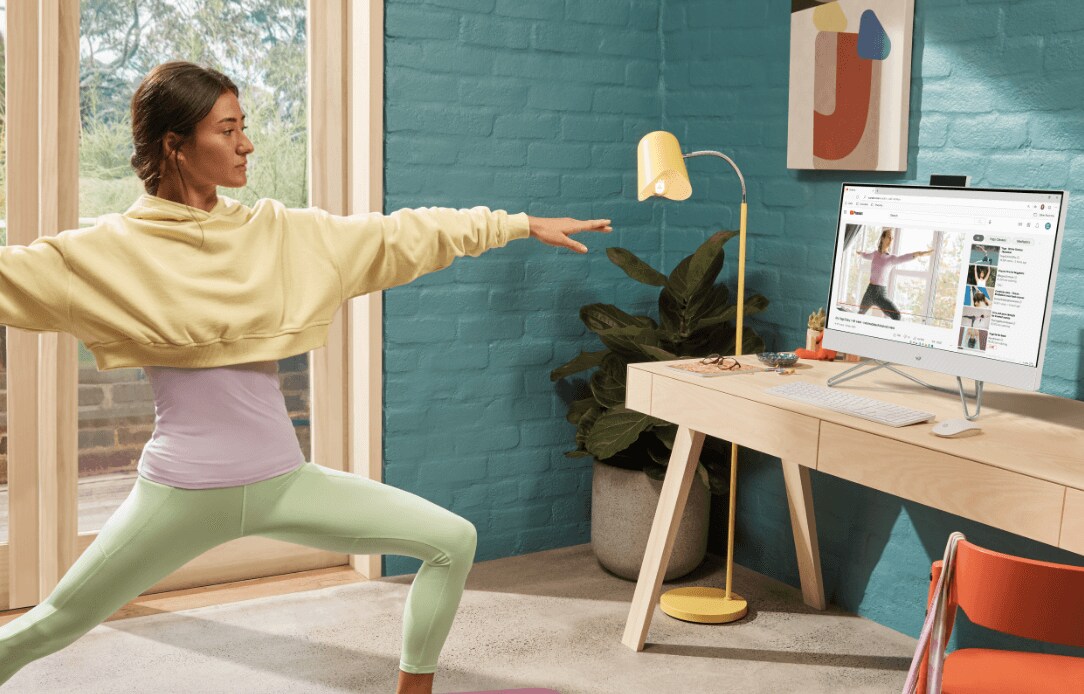 A woman practicing Yoga watching a virtual class on an HP All-in-One PC on top of a wooden table.