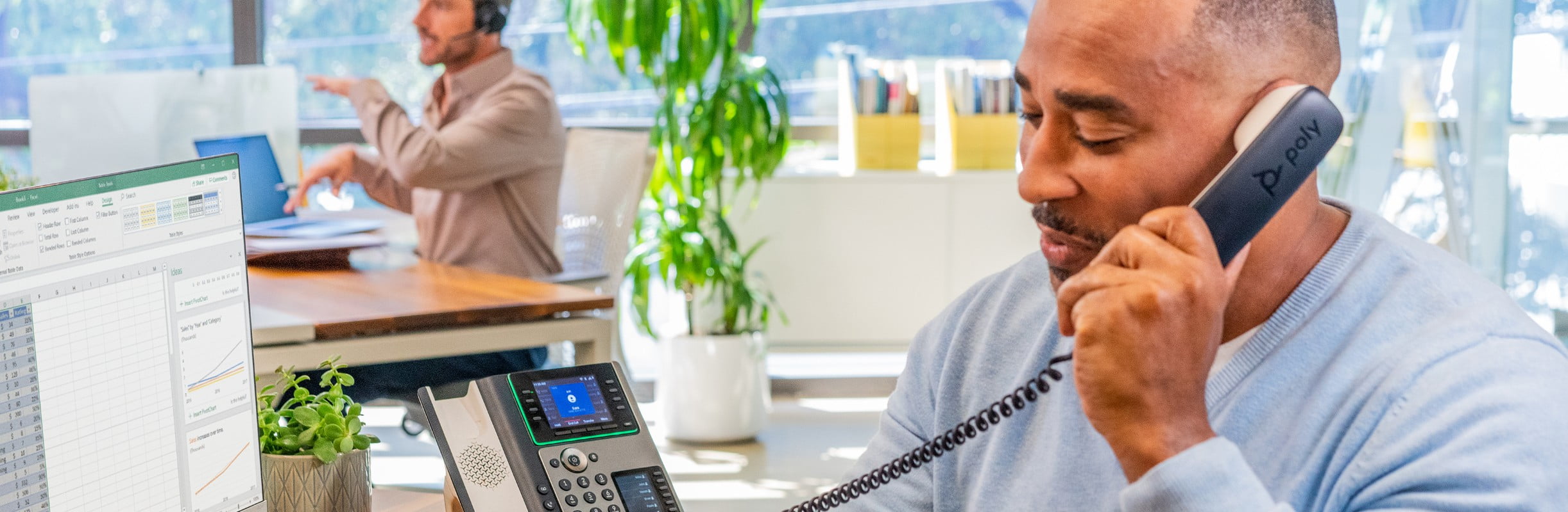 Man seated at desk in an open plan office, engaged in a call using the Poly Edge E450 phone with handset​