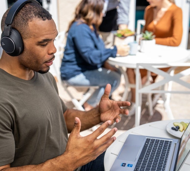 Man sitting outside at café table using Poly Voyager surround 80 headset and HP laptop during a video call