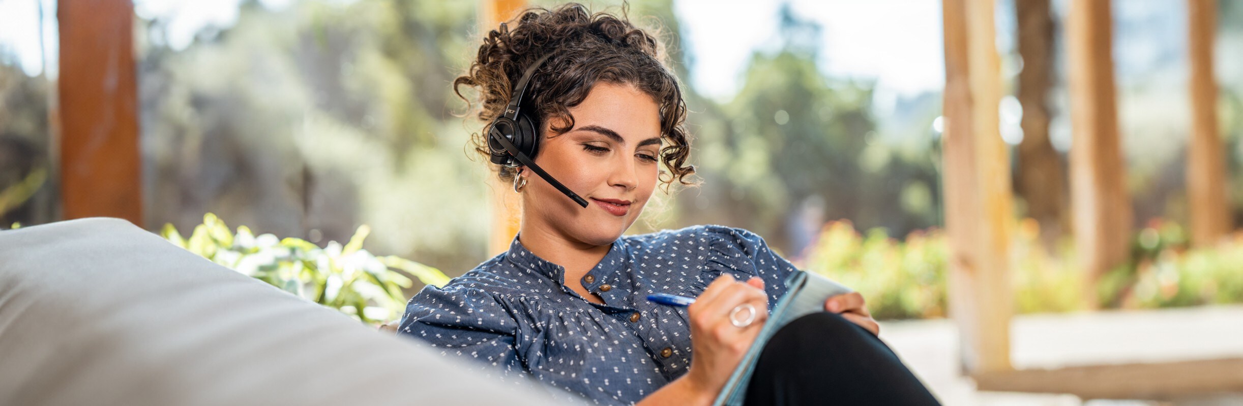 Woman working from home, seated on sofa and writing in notepad while wearing Poly Savi 8210 wireless headset