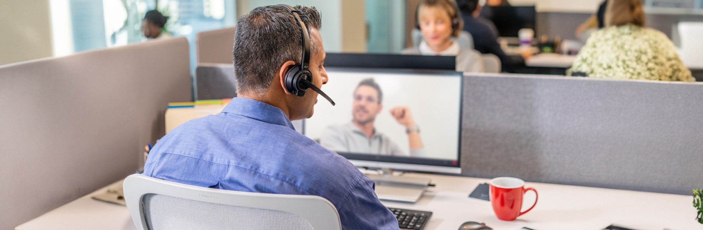 Man seated at desk in open office, talking on video call using Poly Savi 8210 wireless headset