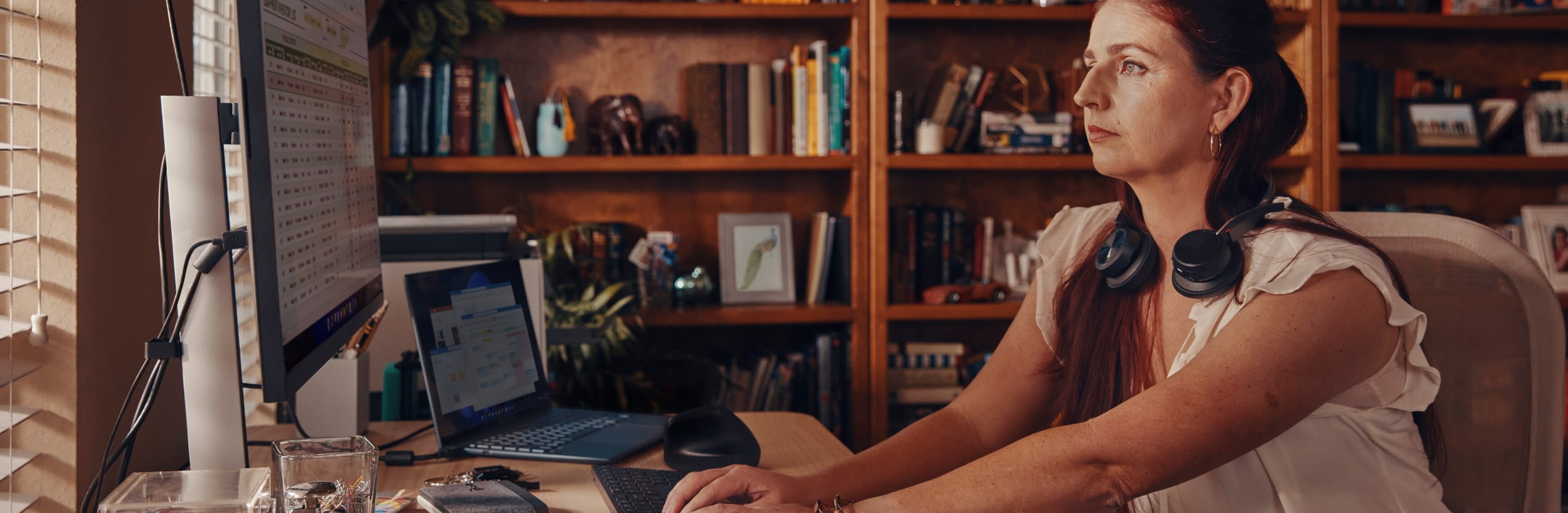 Woman working at home office desk, wearing Poly Voyager Focus 2 stereo headset draped comfortably around her neck while not in use