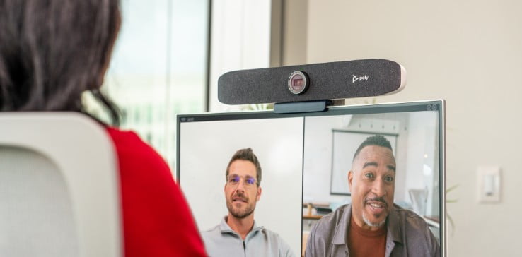 Blick über die Schulter einer Person am Büroschreibtisch, die im Videocall die Poly Studio P15 Videoleiste auf dem Monitor einsetzt