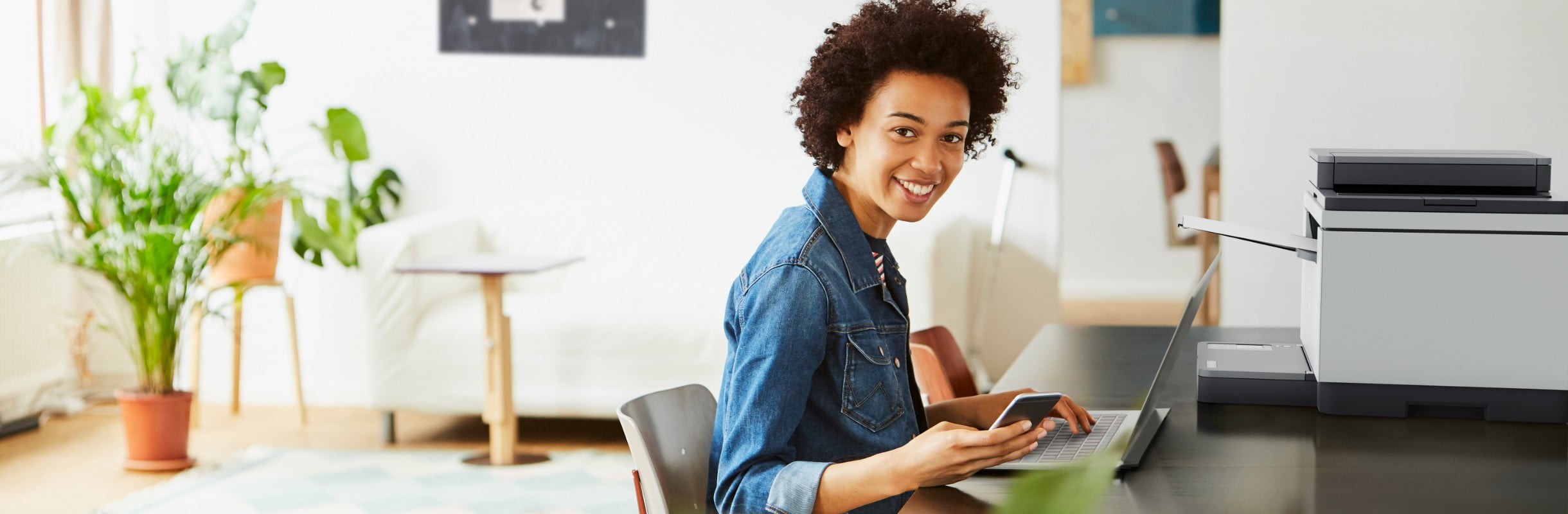 A woman sits at a desk in front of an HP laptop, an HP printer stands next to her, and looks at the phone in her hand.