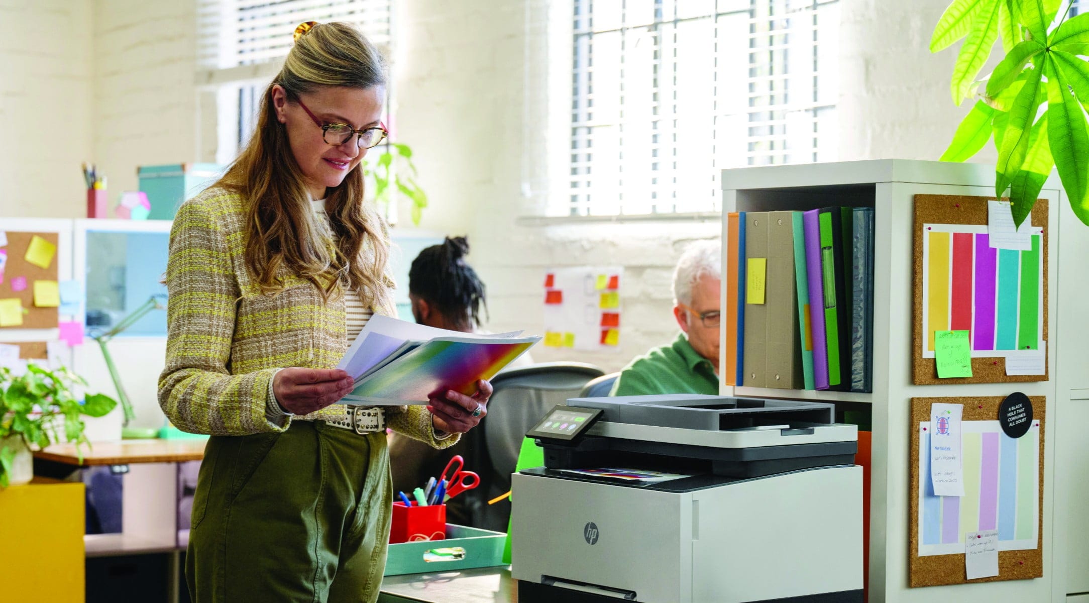 En kvinne som står ved siden av en Laserjet Pro-skriver på kontoret og holder utskrifter i hånden