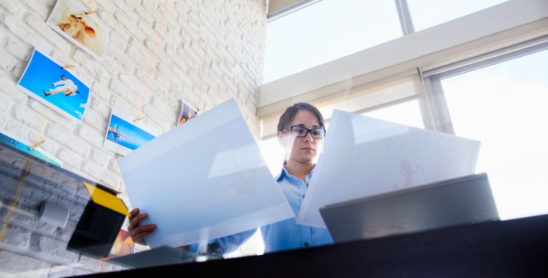 A person sorting through photography printouts in a modern, bright office with photos on the wall and large windows in the background.