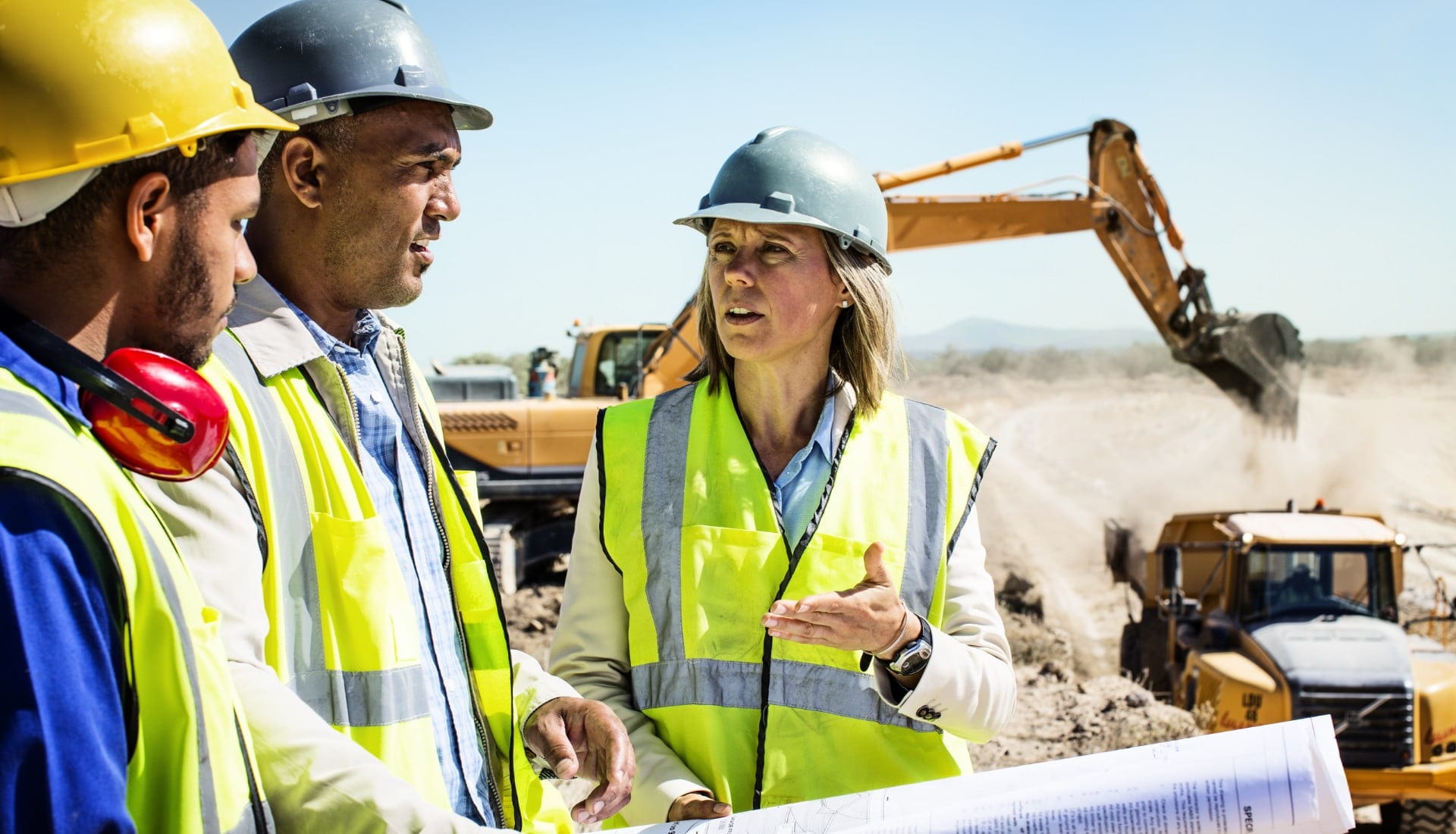 Three construction workers on a worksite with machinery in the background.