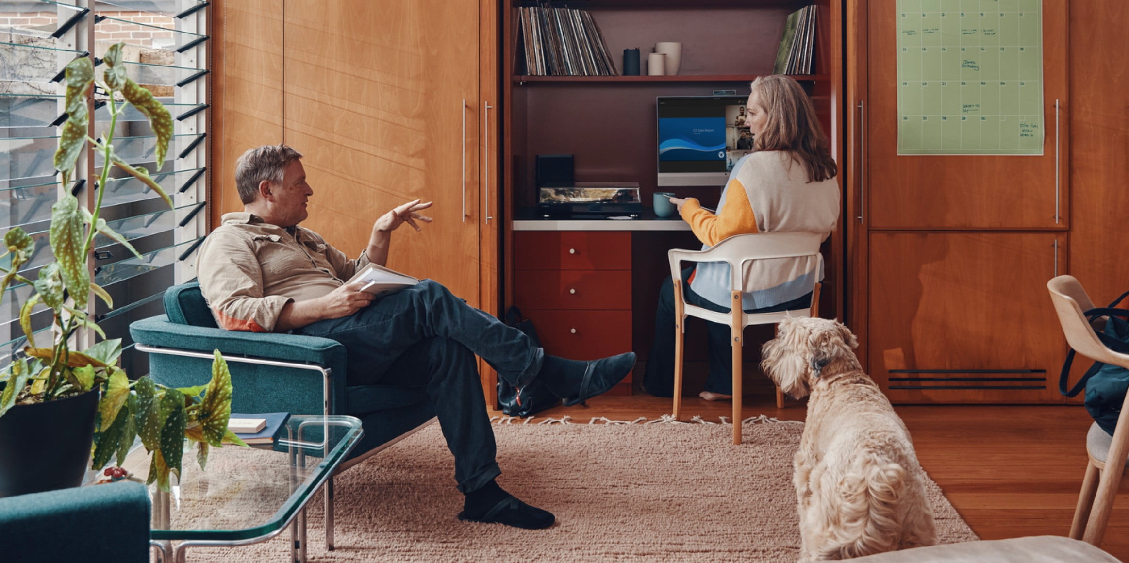 En kvinde arbejder fra sit skrivebord derhjemme, mens hun interagerer med et familiemedlem, der sidder i nærheden.