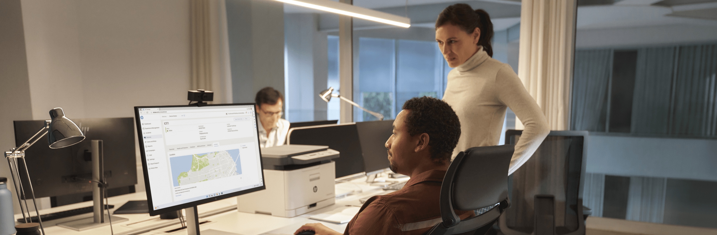 Two coworkers at a desk with an HP laptop, monitor and printer.