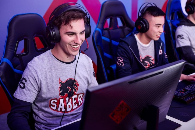 Professional esports player wearing a Saber team jersey laughs while competing at a gaming station, with teammates visible in gaming chairs behind him