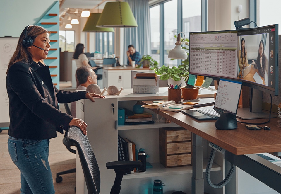 A woman with a headset stands before a computer monitor, focused on her work in a modern office environment.